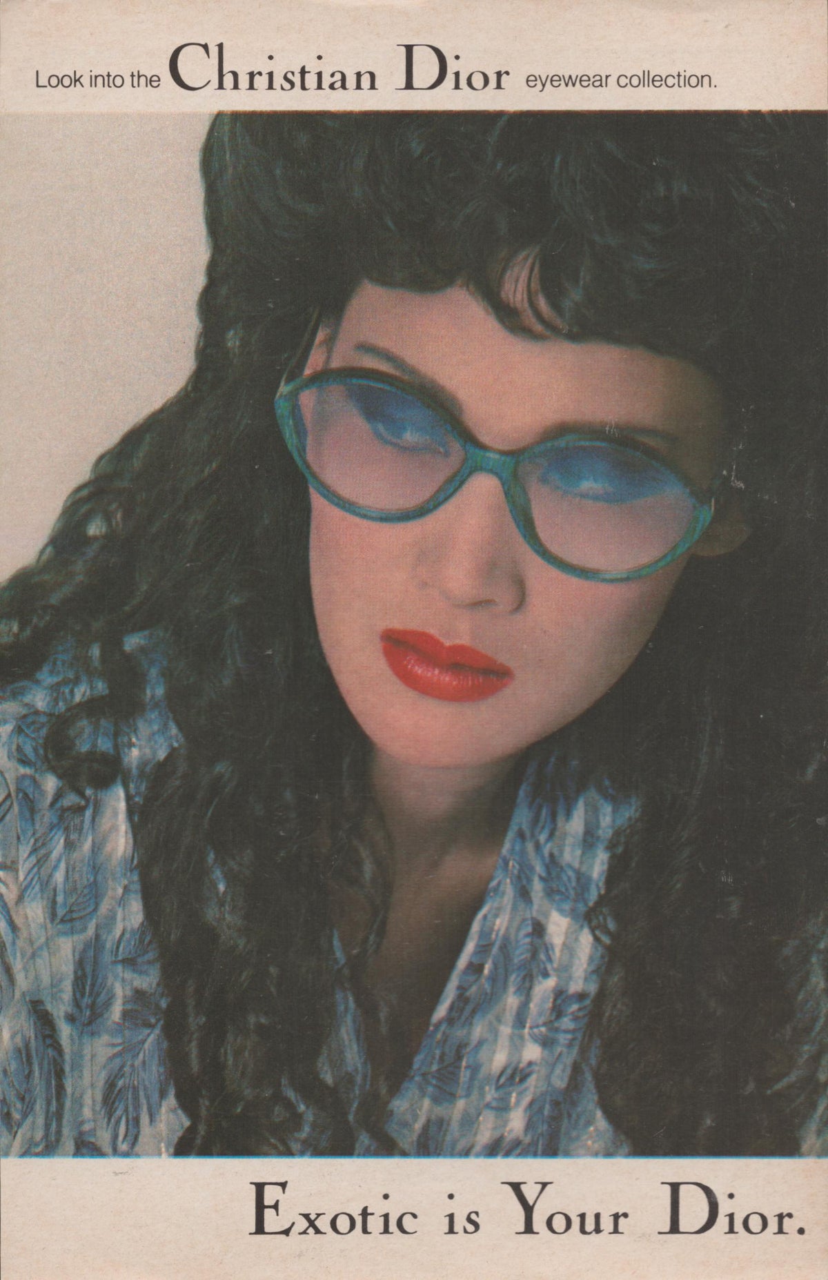 Vintage 1980s Christian Dior Eyewear original print advertisement, close-up portrait of dark-haired model wearing oversized teal sunglasses with red lips and tropical print blouse, 'Exotic is Your Dior' headline