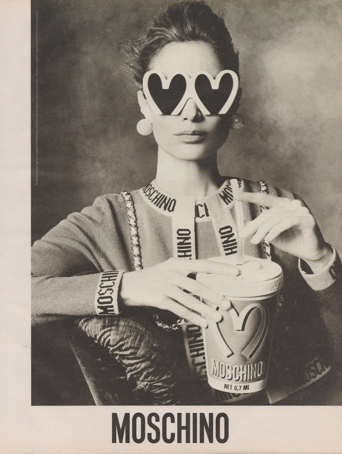 Vintage 1990s Moschino original print advertisement, black and white image of model in logo-band knit wearing heart-shaped sunglasses holding a Moschino-branded fast food cup