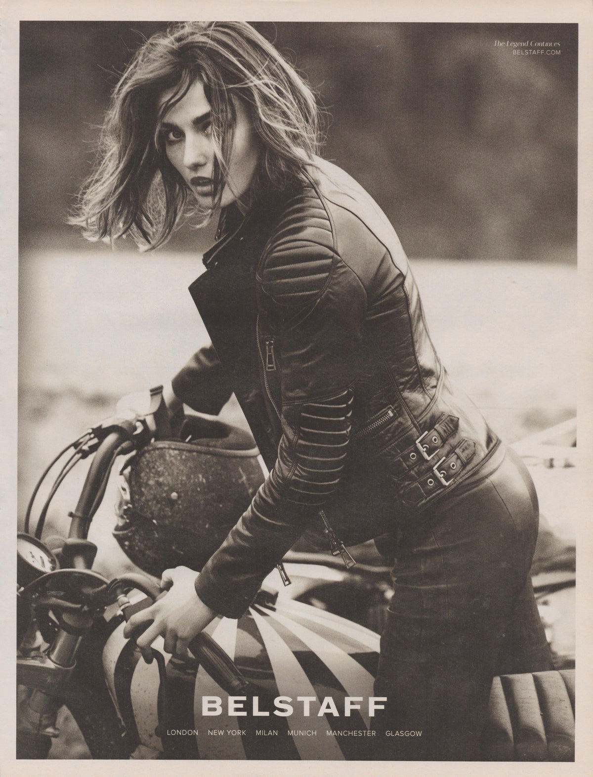 Vintage 2000s Belstaff leather motorcycle jacket original print advertisement, sepia-toned image of wind-blown female model astride a cafe racer motorcycle