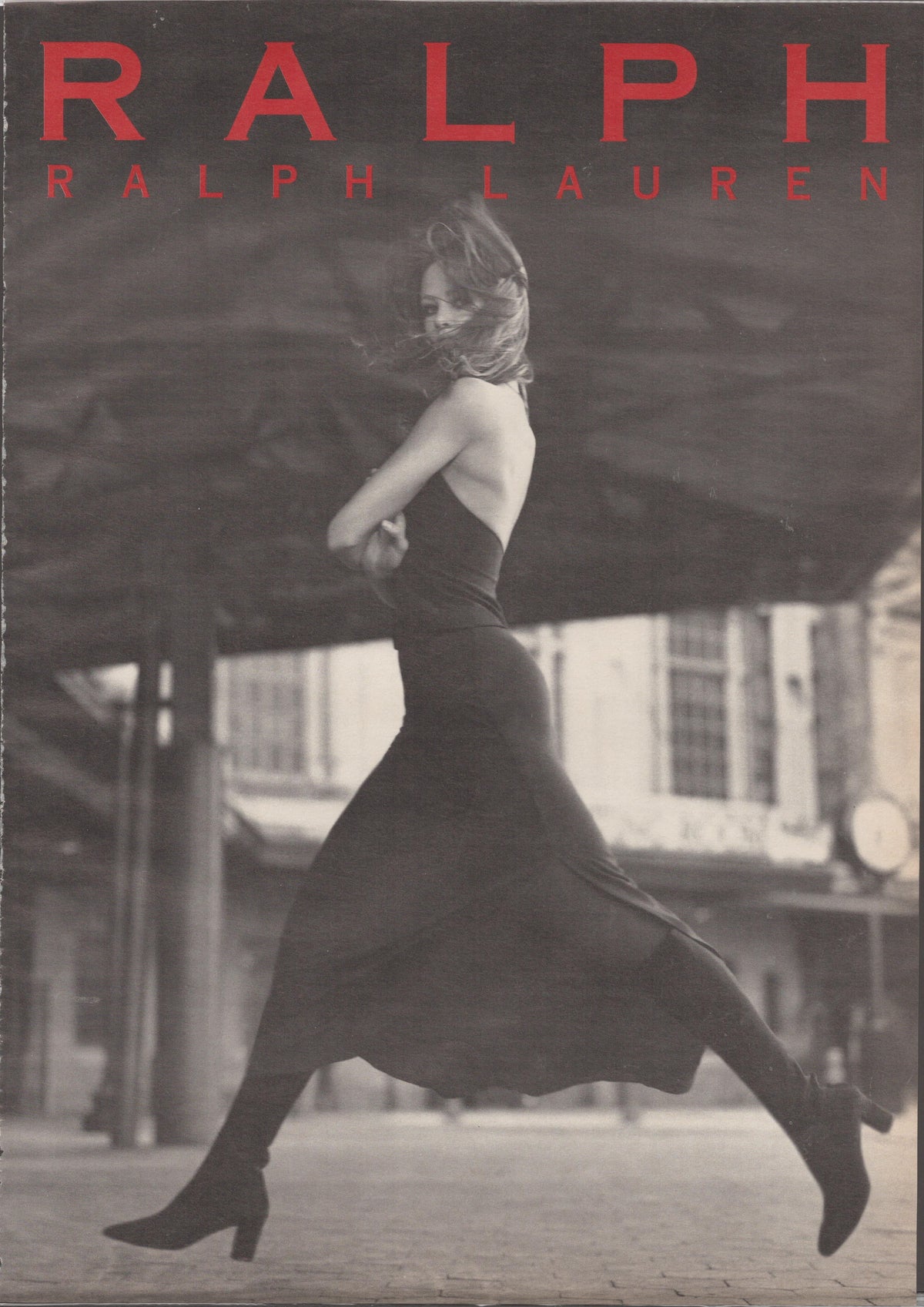 Vintage 1990s Ralph Lauren 'Ralph' fragrance original print advertisement, black and white photo of woman leaping mid-stride on cobblestone street in halter dress and heeled boots with bold red Ralph Lauren typography