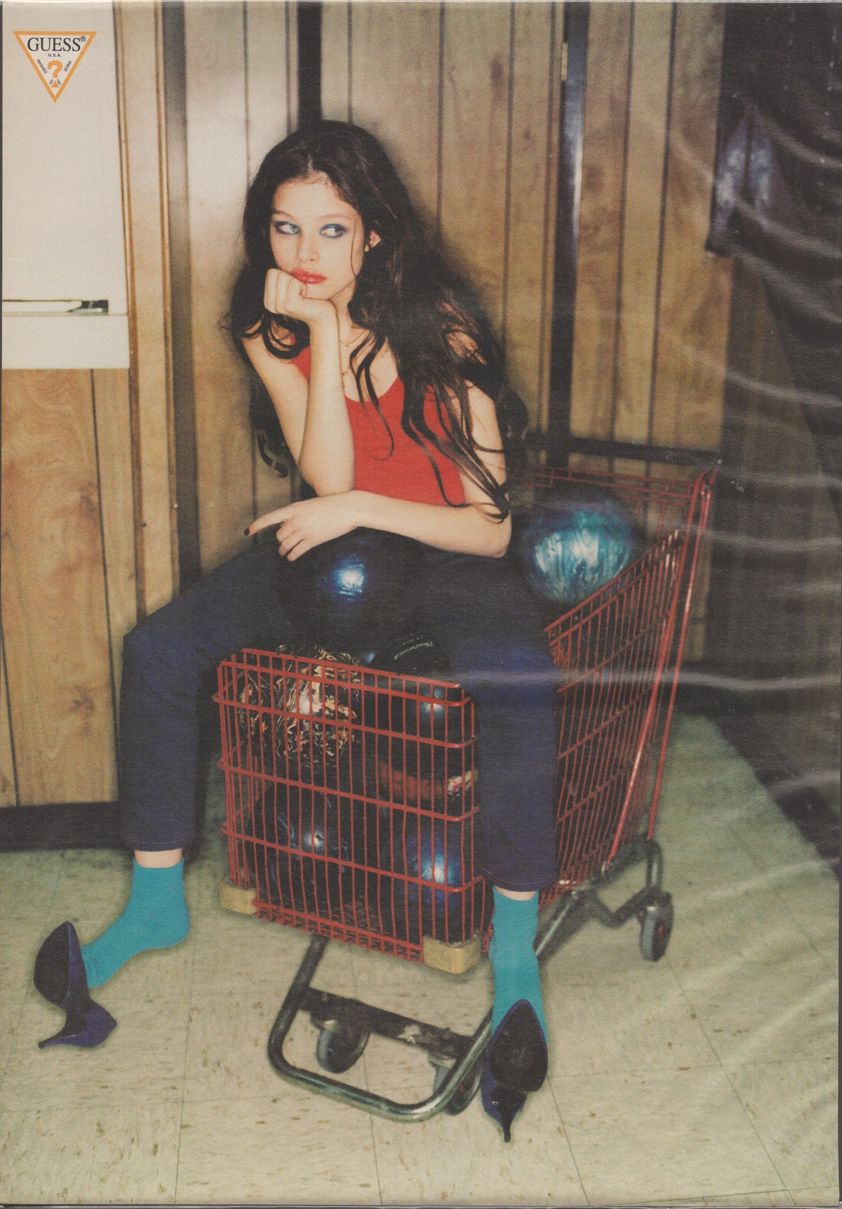 Vintage 1990s Guess Jeans original print advertisement, dark-haired model seated in red shopping cart wearing slim jeans, red tank top, teal socks, and heels against wood-paneled backdrop
