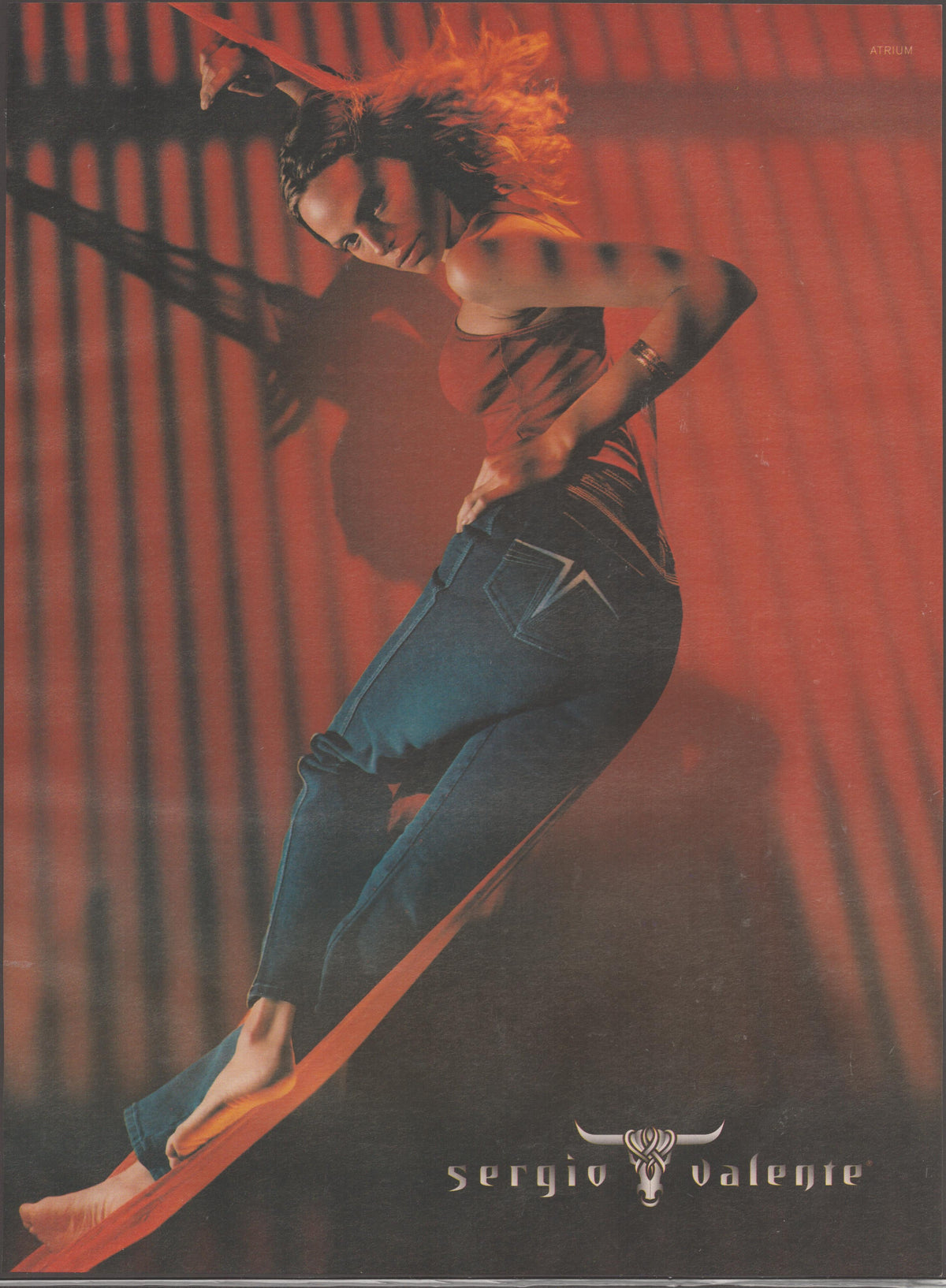 Vintage 1980s Sergio Valente jeans original print advertisement, female model posing dynamically against a red background wearing signature slim-cut denim jeans with bull-skull logo