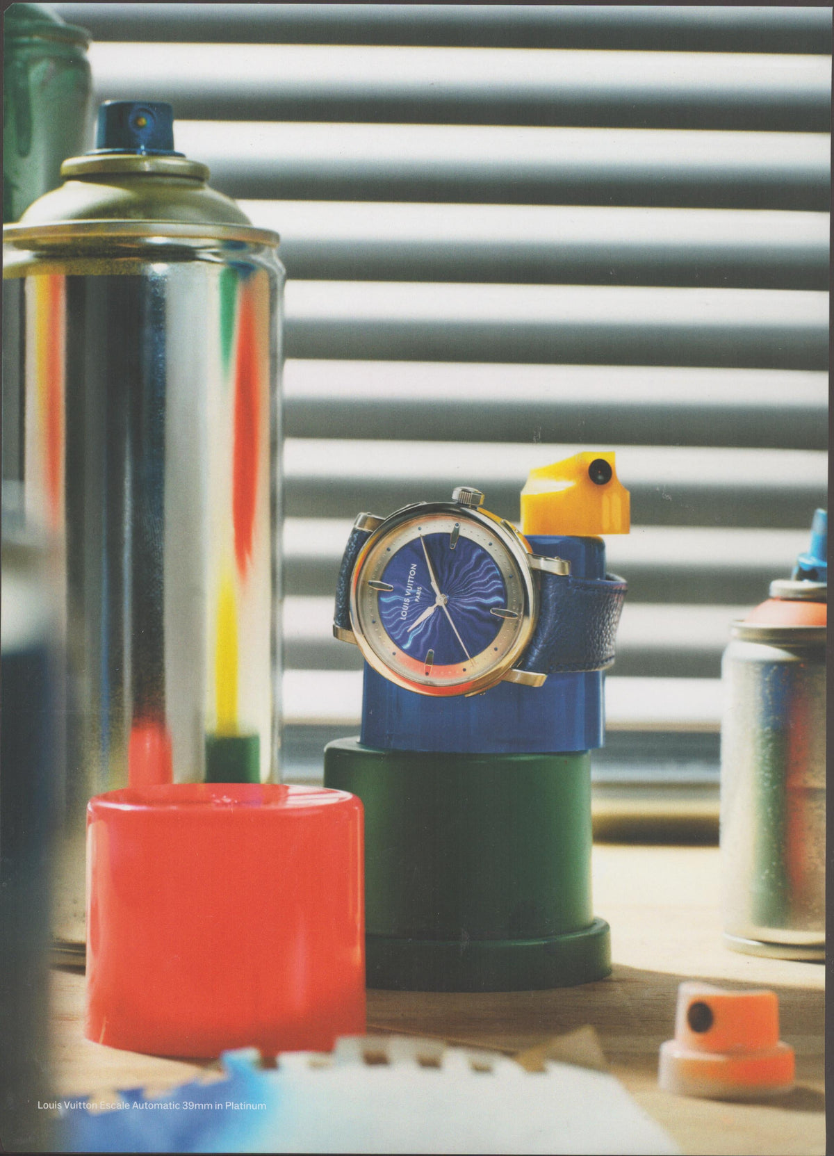 Vintage Louis Vuitton Escale Automatic 39mm Platinum original print advertisement, blue dial watch displayed among colorful spray paint cans and geometric props on a wooden surface