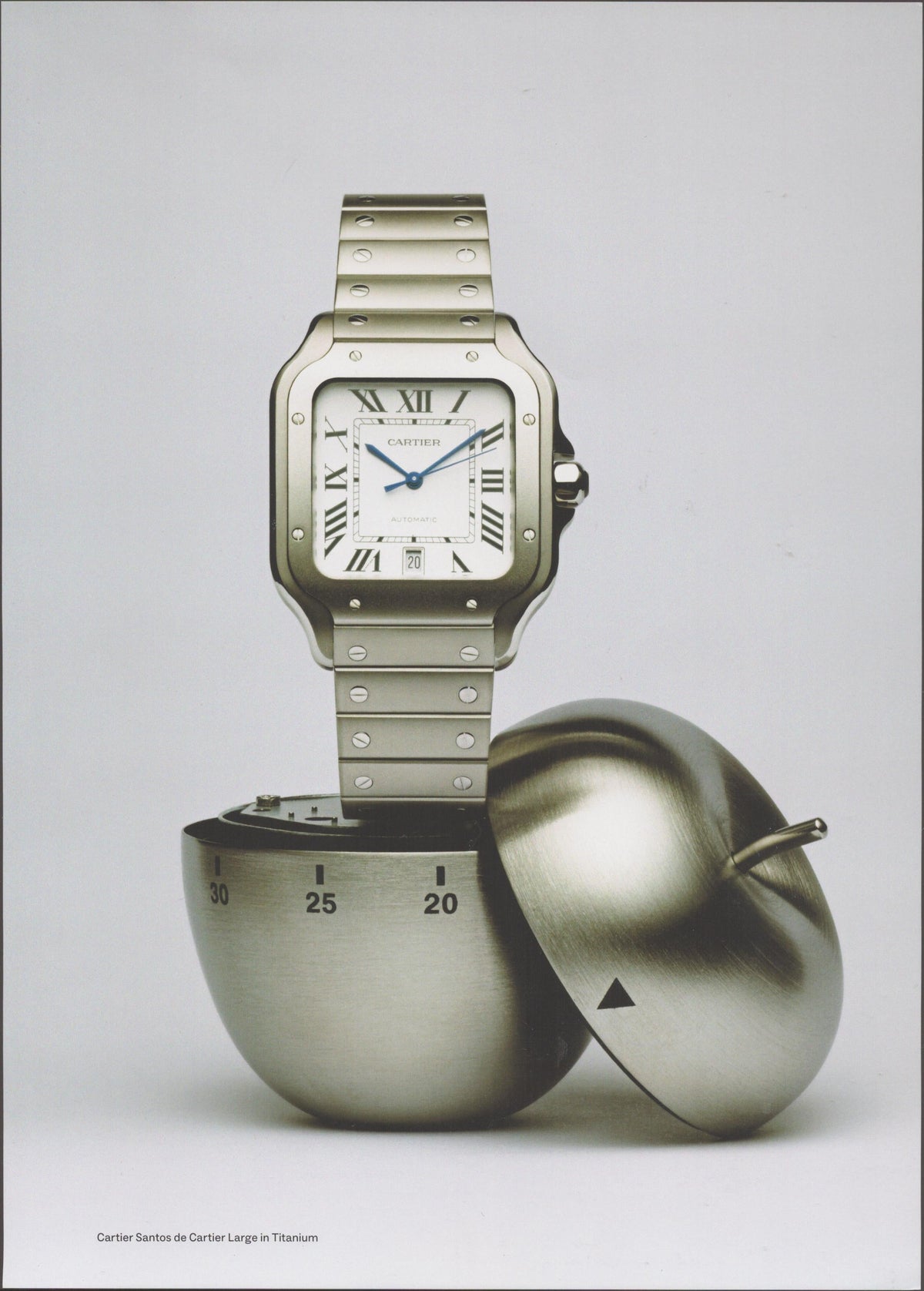 Vintage 2010s Cartier Santos de Cartier Large Titanium original print advertisement, square titanium watch balanced atop a sculptural titanium apple timer on neutral gray background