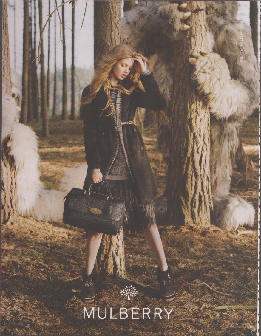 Vintage 2000s Mulberry original print advertisement, model in autumn layers holding black leather satchel in an enchanted forest setting with large furry creatures among pine trees