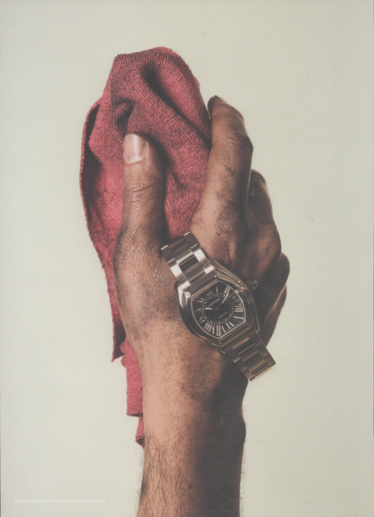 Vintage 2002 Cartier Roadster original print advertisement, close-up of a grease-stained hand gripping a red shop rag with a stainless steel Cartier Roadster watch on the wrist against a neutral background