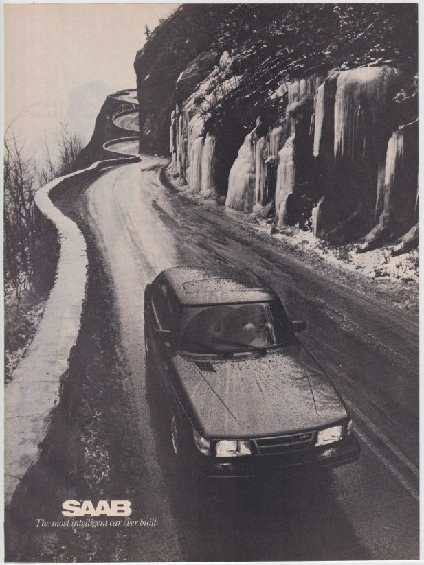 Vintage 1980s Saab 900 original print advertisement, black and white aerial view of Saab navigating icy mountain road with frozen waterfalls on rocky cliff face