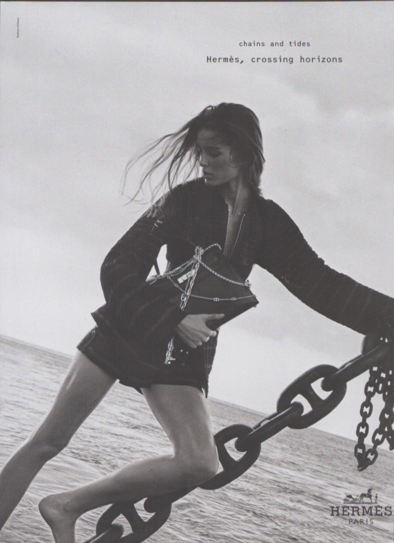 Vintage 1990s Hermès Paris original print advertisement, black and white image of windswept model holding chain-detailed handbag against dramatic ocean and nautical chain backdrop